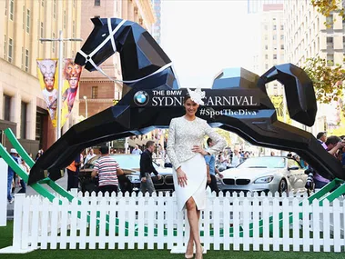 Rachael Finch at BMW Sydney Carnival, poses in front of a geometric horse sculpture, wearing a white outfit and fascinator.