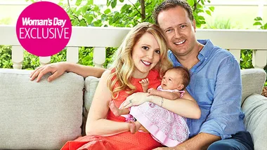 A smiling couple sits on a couch, holding a baby in a pink dress, with 'Woman's Day Exclusive' logo at the top left.