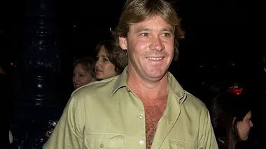 Steve Irwin smiling and walking outdoors, wearing a beige shirt, with people and a lamppost in the background.