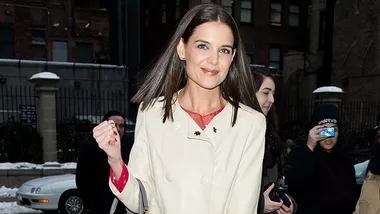 Katie Holmes smiling and wearing a cream coat outdoors, with people and a snowy street in the background.