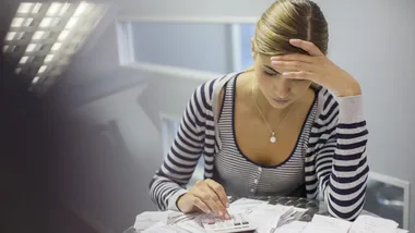 Woman calculating expenses with a stressed expression, surrounded by bills and receipts at a desk.