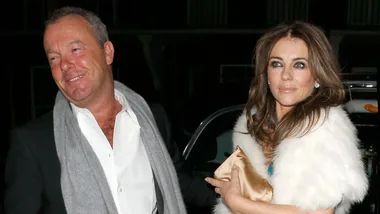 A man in a suit with a scarf and a woman in a white fur stole walk together at night, both smiling slightly.