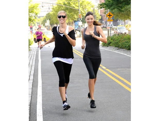 Two women jogging on a road, wearing black athletic outfits, with trees and vehicles in the background.