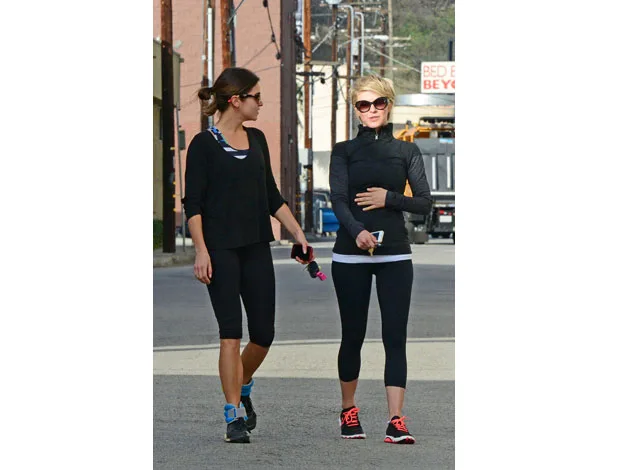 Two women in athletic wear walking on a street, one holding a phone and keys, near an industrial backdrop.
