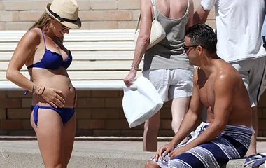 Pregnant woman in a blue bikini touching her baby bump, talking to a man in swim trunks by a pool.