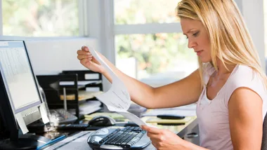 A woman working at a desk, reviewing a document, in front of a computer in a well-lit office space.