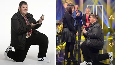 Man in a checkered shirt kneeling with a ring, proposing on a TV show stage to a woman dressed in black.