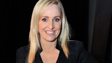 Blonde woman smiling, wearing a black jacket, against a dark background.