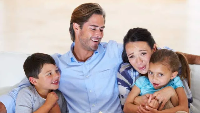 A happy family of four sits on a couch, laughing and hugging, in a bright living room.