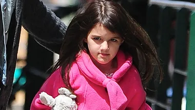 Young girl in a pink coat holding a stuffed toy, walking outdoors with an adult.