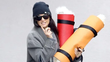 Woman in sunglasses and a beanie carrying two yoga mats, one red and one orange.