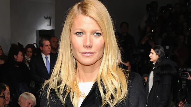 Gwyneth Paltrow at an event, wearing a black coat, surrounded by people and photographers in the background.