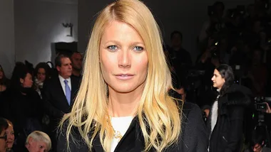 Gwyneth Paltrow at an event, wearing a black coat, surrounded by people and photographers in the background.