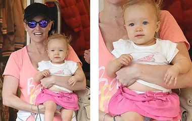 Mom in sunglasses holding a baby in a pink skirt, both smiling, during an outing.