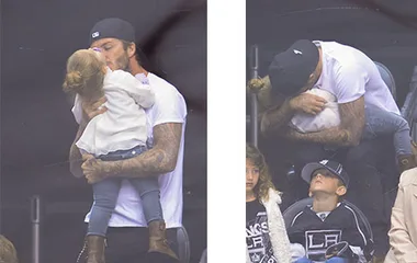 Father in cap lovingly kisses daughter who is wearing a white sweater while at a sports event.