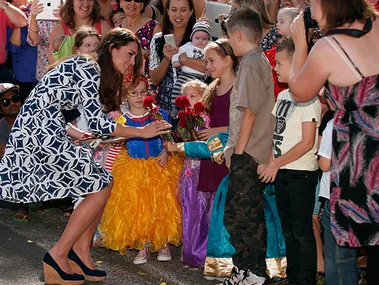 Kate Middleton greets children in colorful costumes, accepting flowers, while surrounded by an enthusiastic crowd.