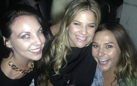 Three women smiling at a social event, one holding a drink, all dressed casually.