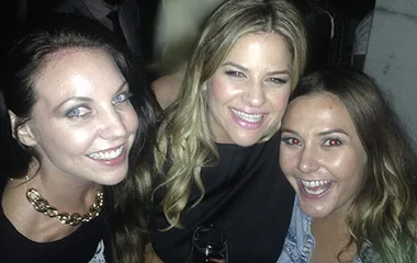 Three women smiling at a social event, one holding a drink, all dressed casually.