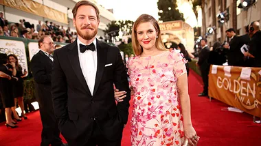 Couple posing on the red carpet at the Golden Globe Awards, woman in a floral dress and man in a tuxedo, smiling to the camera.