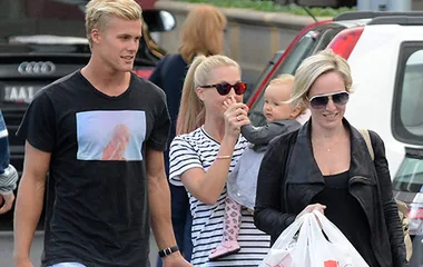 Group of adults walking outdoors; woman holds a baby while another carries shopping bags. Casual attire, daytime setting.
