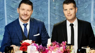 Two well-dressed men seated at a table with colorful flowers, against a textured blue backdrop.