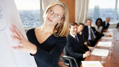 A woman presenting at a business meeting with colleagues seated at a conference table in the background.