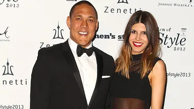 Man in a tuxedo and woman in a black dress smiling at a formal event backdrop with logos.