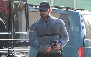 Bradley Cooper in a gray workout shirt and cap, holding gym items near vehicles, preparing for a role.