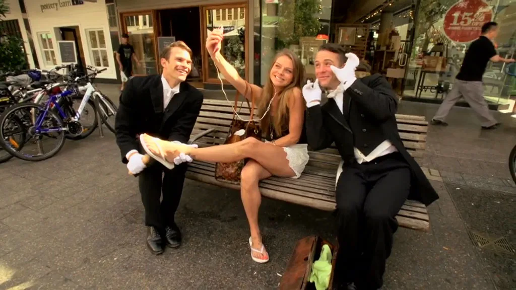 Two men in butler uniforms and a woman taking a selfie on a bench in a bustling street.