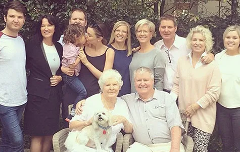 Family gathering with adults and a child. An elderly couple seated with a white dog in front. Lush greenery in the background.