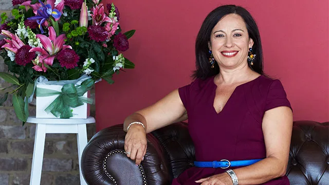 Woman in a burgundy dress smiles, sitting on a leather couch with a large flower arrangement beside her.