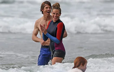 Two people in wetsuits joyfully playing in ocean waves, with a child in foreground.