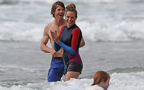 Two people in wetsuits joyfully playing in ocean waves, with a child in foreground.