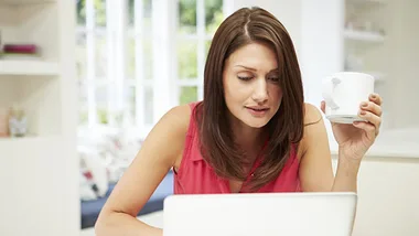 Woman working on a laptop at home, holding a coffee mug.