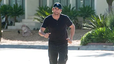 A person in a black shirt and cap walking outdoors on a sunny day, surrounded by greenery and a building in the background.