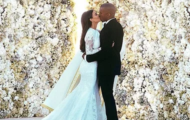 Couple kissing in front of a floral wall, bride in lace gown, groom in black suit, capturing a wedding moment.