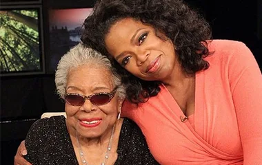 Two women seated together smiling, one wearing sunglasses, the other with her arm around her.