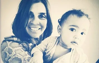 A woman smiling broadly holds a toddler gently, both appearing cheerful in a close-up, warmly lit photograph.