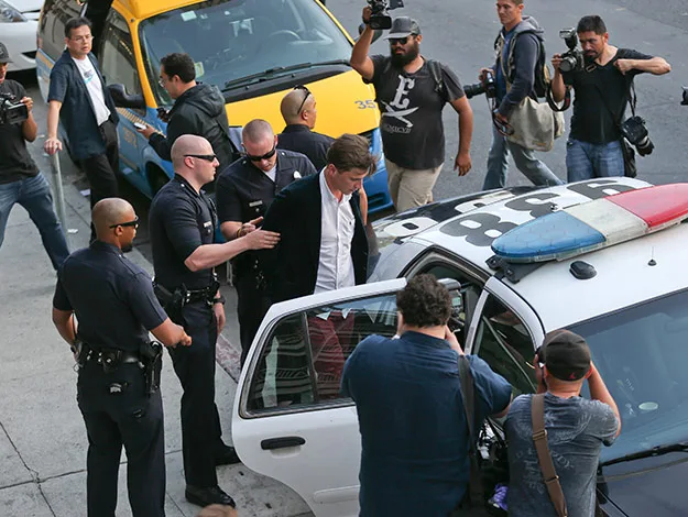 Police arrest a person while photographers and reporters document the scene outside, near a taxi and police car.