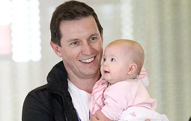 Man holding a baby girl while smiling, in an indoor setting with soft lighting in the background.