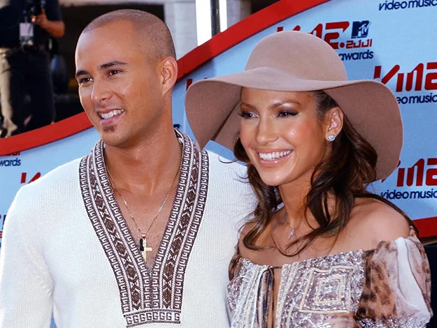 A man and a woman smiling at the MTV VMA red carpet, the woman wears a wide-brimmed hat and patterned dress.