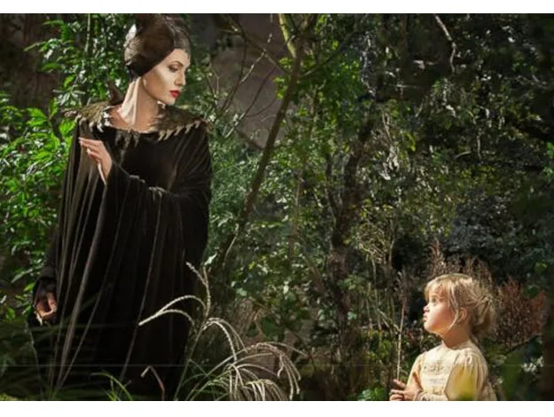Maleficent in dark robes looking at a young girl in a forest, surrounded by lush greenery.