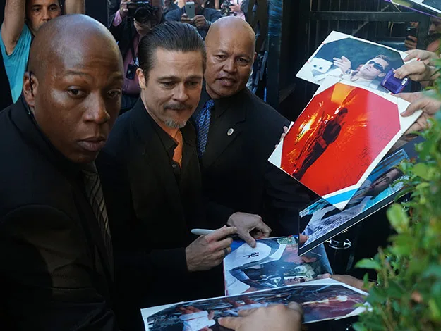 Actor signing autographs for fans, surrounded by security and photographers.