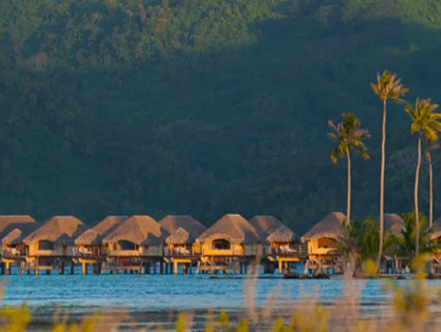 Overwater bungalows on blue ocean with lush green hills and palm trees in the background.