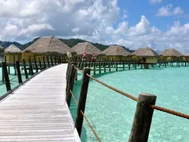Overwater bungalows and wooden walkway above clear blue waters, with a backdrop of mountains and clouds in the sky.
