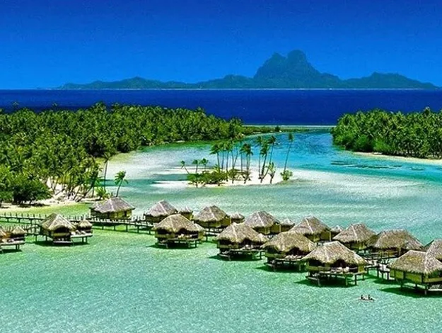 Overwater bungalows on turquoise water with lush green palms and a mountain in the background under a clear blue sky.