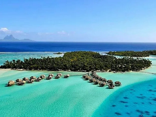Overwater bungalows on turquoise water with lush island and distant mountains under a clear blue sky.