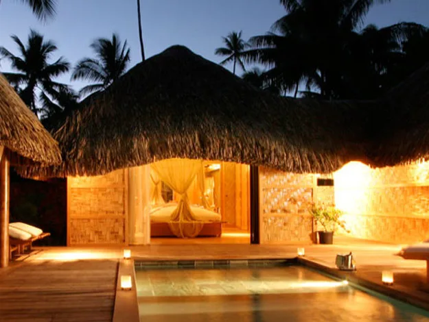 Tropical bungalow with thatched roof, glowing warmly at dusk, overlooking a serene pool surrounded by palm trees.