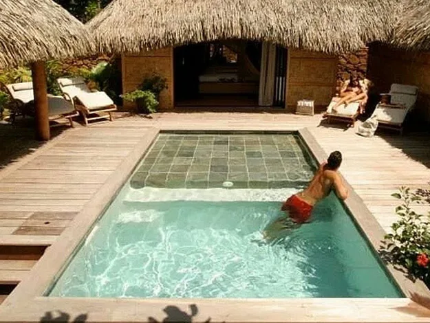 Swimming pool with a man in red shorts, surrounded by wooden deck, thatched huts, and lounge chairs.