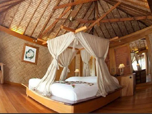 A cozy tropical bedroom with a canopy bed, wooden floors, thatched ceiling, and warm lighting.